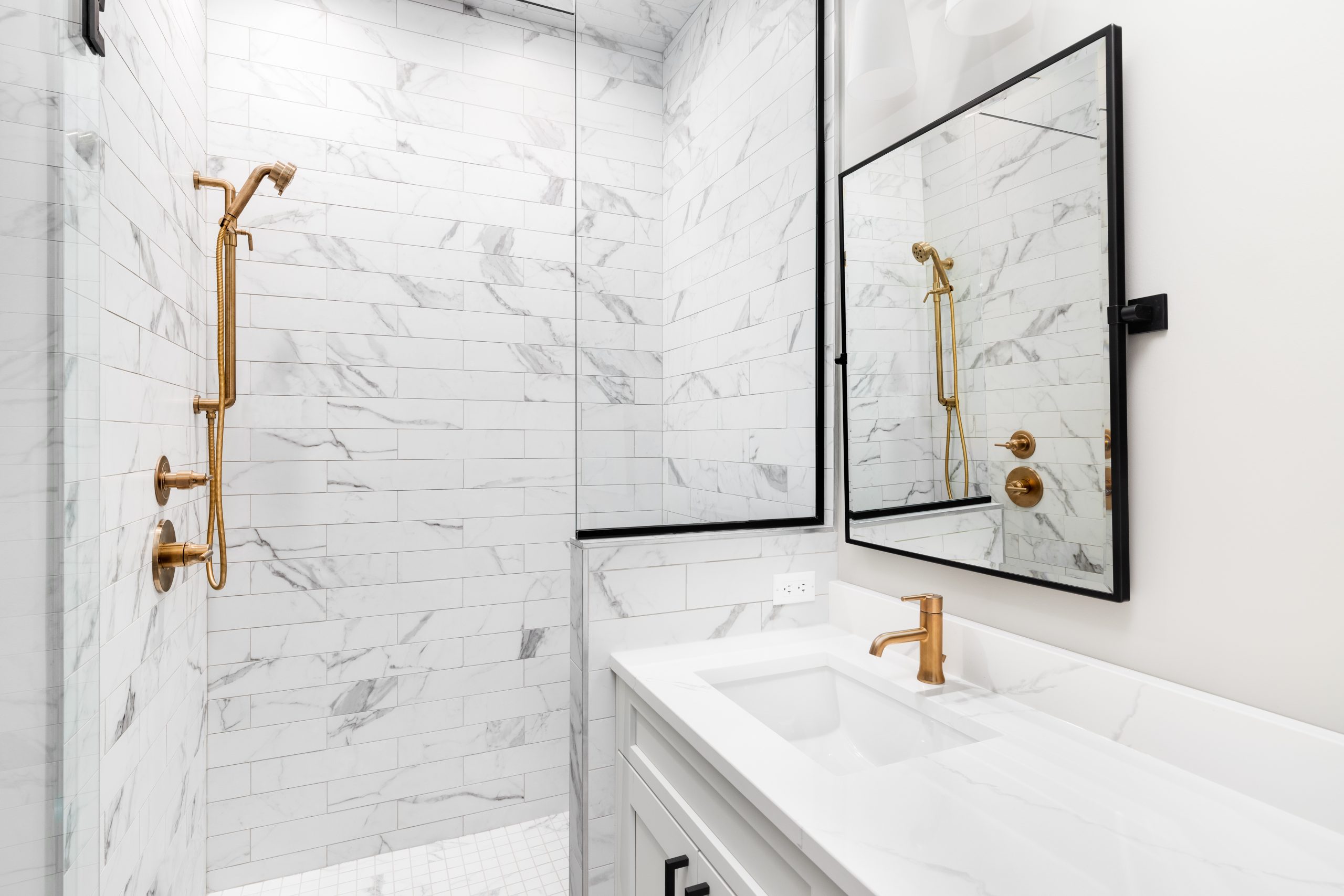 A white bathroom with a marble countertop, gold faucets, black mirror, and a shower with marble subway tiles. No brands or labels. Granite and stone Bathroom restoration